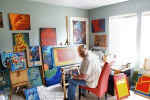 This was the old loft studio that was used around 2012 showing the artist posing at the easel. Seated on a red chair borrowed from the dining table downstairs. The move to the upstairs loft was enabled when his adult children moved away from home. This left space which could be converted into a painting studio. The painting space prior to this had been a small corner carved out in a corner of the kitchen eating nook where it had been for 3 years before. That red kitchen chair was a reminder of those years. Also surrounding the artist are the creations from the 'In Red' and 'Human Security' series. In the background on the door is the blow up for the digital mock up of the partially done painting (on the floor against the wall) 'In the Beginning.' Photo taken May 2012