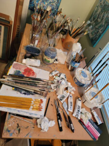The work table of the artist (as shown from the days of the old north window studio). Not one for much fancy 'store bought' equipment, the table top is a scrap of pressed board on a discarded wood table that was given him from his last employer. Old pickle jars for brushes. Golden heavy body acrylics are the standard paint in tubes used for most all work done since about 2013. Photo taken September 2021